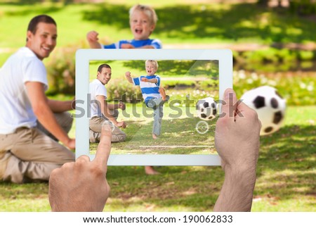 Hand holding tablet pc showing father watching son kicking football