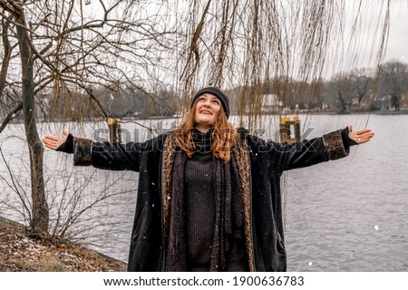 young brunette woman standing on snowy day by lake and spreading her arms
