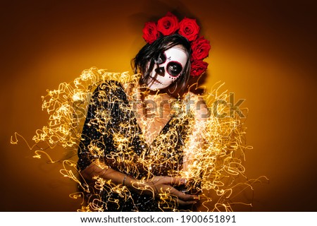 Mysterious lady with crown of flowers surrounded by shining garlands. Portrait of Mexican girl with skull-shaped make-up