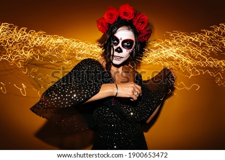 Adult woman with unusual makeup for Halloween spends time at party. Dark eyed model in black top mysteriously looking into camera