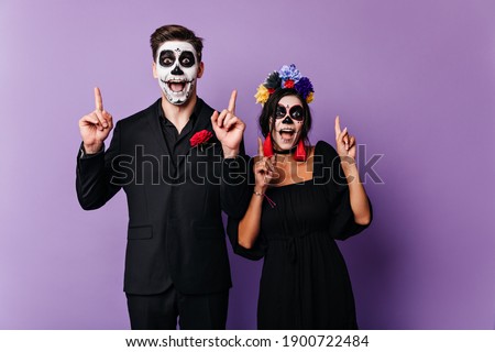 Cheerful dark-eyed boy and girl emotionally pose, showing fingers up. Shot of couple with face art in Mexican style on purple background