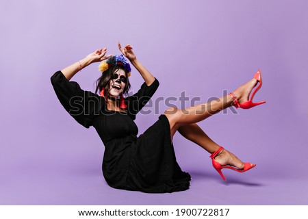 Curly brunette with face art for Halloween sings while sitting on floor. Photo of girl in high spirits on lilac background