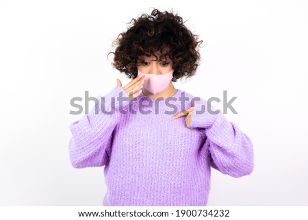 Shocked young beautiful caucasian woman wearing medical mask standing against white wall look surprisedly down, indicates at blank  with fore finger, Scared model