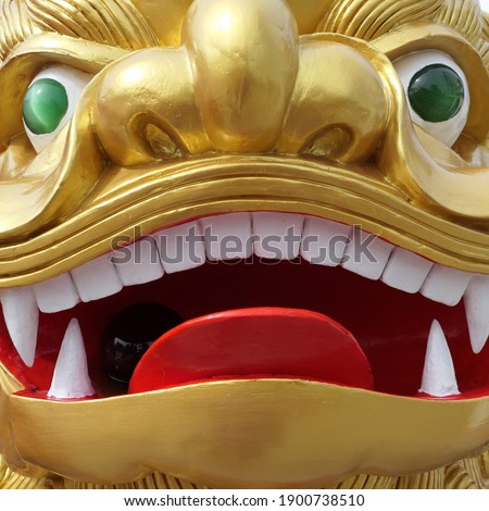Phuket, Thailand, October 02, 2018: Closeup square picture of a gold-painted Singha or lion statue with green eyes, red tongue, and bright white teeth.