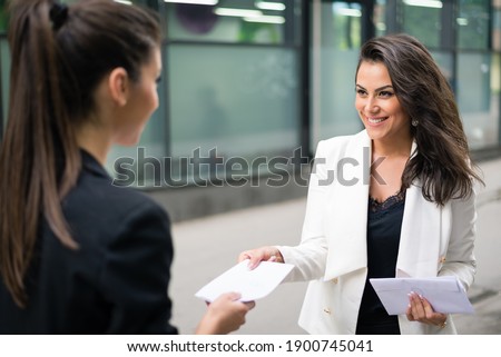 Two business women sharing documents in front of modern corporate building. Female coworkers handling paperwork Royalty-Free Stock Photo #1900745041