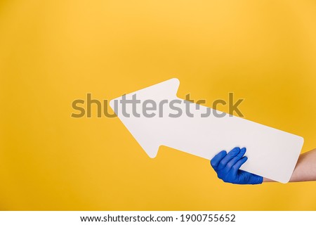 Close up of unrecognizable female in medical protective gloves hold white paper arrow pointing at copy space for advertisement, isolated on orange studio wall. Advertising area, mockup. Hand gesture