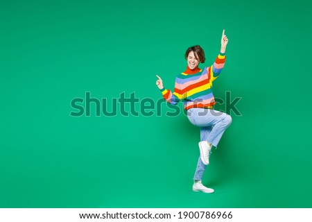 Full length side view of cheerful funny young brunette woman 20s in casual colorful sweater dancing pointing index fingers up winner gesture isolated on bright green color background studio portrait