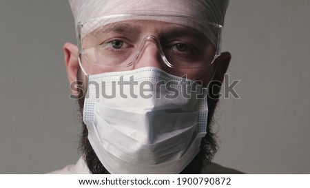 Portrait of a tired middle-aged doctor with a mask on his beard, wearing personal protective equipment on a gray background. Concept of the coronavirus pandemic.