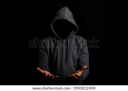 Man without a face in a hood holds something in his hands on a dark background. Royalty-Free Stock Photo #1900822498
