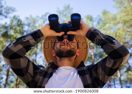 Men tourist in a hat and a gray checkered shirt looks through binoculars on a forest background.