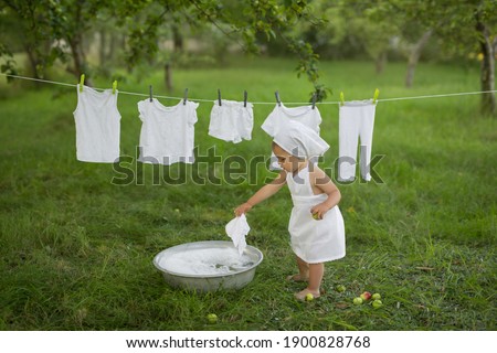 A child cheerfully laundered his clothes in the basin. Girl in white dress hanging wet laundry to dry. Washing in a vintage basin. Hand wash. soap powder. Laundry conditioner. Snow-white underwear