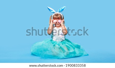 A little girl with white hair and bunny ears on her head sits on a blue background and closes her eyes with two Easter eggs. Concept of holiday - Easter. Christ is risen. Banner. A place for text.