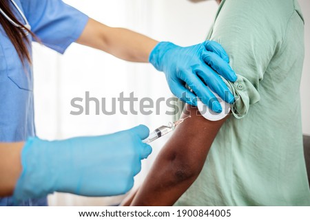 Female doctor wearing blue latex gloves injecting a woman in her arm with a needle and syringe containing a dose of the COVID-19 vaccine cure by way of immunisation Royalty-Free Stock Photo #1900844005