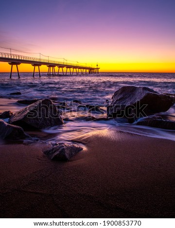 Picture of the sun, the waves and the rocks of the Badalona beach with the Pont del Petroli behind during sunrise.