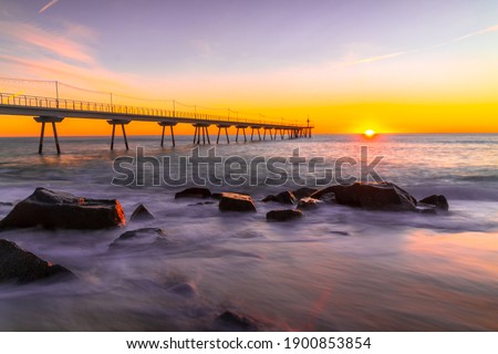 Picture of the sun, the waves and the rocks of the Badalona beach with the Pont del Petroli behind during sunrise.