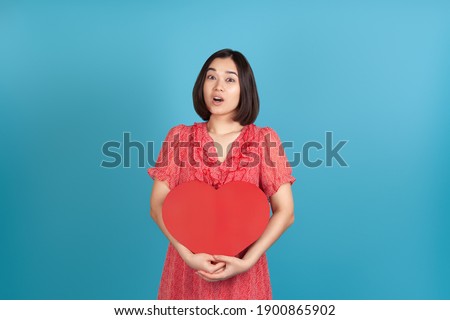surprised, delighted, excited young asian woman with open mouth in red dress holding big red paper heart isolated on blue background.