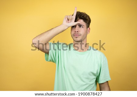 Handsome man wearing a green casual t-shirt over yellow background making fun of people with fingers on forehead doing loser gesture mocking and insulting.