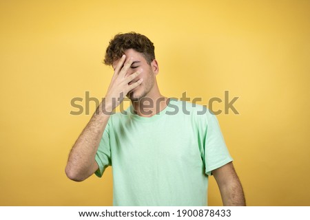 Handsome man wearing a green casual t-shirt over yellow background peeking in shock covering face and eyes with hand, looking through fingers with embarrassed expression