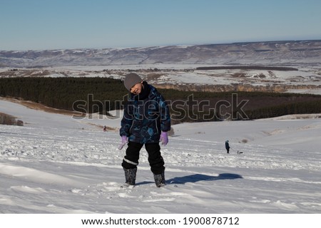 A boy runs across a snow-covered field, mountains and blue sky are visible in the background