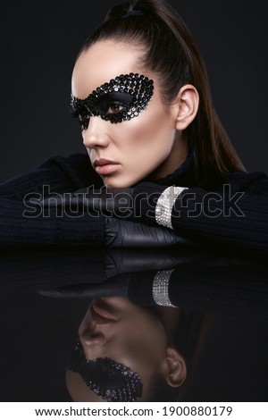 Portrait of charming elegant brunette woman in black turtleneck sweater and sequins mask posing on a mirrored surface in studio