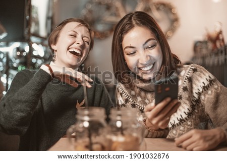 Two funny female friends look at new memes on social networks and laugh out loud in cafe. Smartphone with online Internet access - a new type of leisure for group of friends Royalty-Free Stock Photo #1901032876