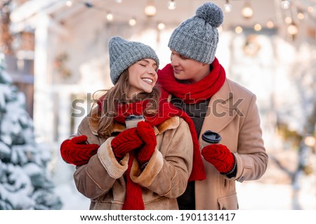 Loving beautiful couplein warm outfits with coffee to go enjoying snowy and sunny winter day, handsome man hugging his happy woman, drinking hot tea while walking outdoors, copy space