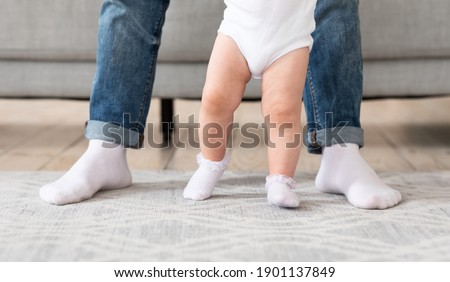 Father Helping His Baby Toddler Make First Steps Learning To Walk Standing Together At Home. Dad's And Kids Legs. Fatherhood And Cute Family Moments Concept. Cropped