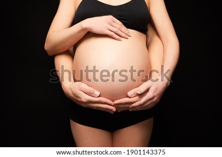 Pregnant woman and her husband's hands hugging the tummy isolated on black background.Portrait of a young couple waiting for a baby. 