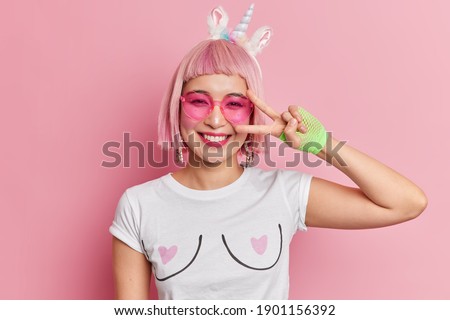 Headshot of cheerful young Asian woman with pink hairstyle smiles pleasantly makes victory gesture wears trendy shades and casual t shirt isolated over rosy background. Body language concept