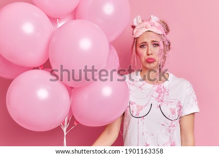 Photo of frustrated displeased blonde woman looks sadly at camera smeared with cream wears blindfold and casual t shirt feels unhappy at party holds helium balloons isolated over pink background