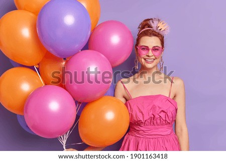 Cheerful sincere woman from ninenties dressed in fashionable pink outfit holds colorful inflated balloons has fun at bridal shower smiles gladfully isolated over purple background. Celebration concept
