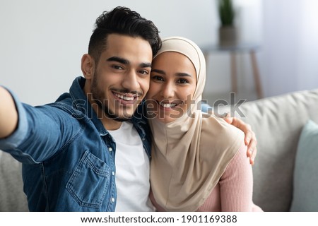 Beautiful muslim man and woman in hijab taking selfie from home, copy space. Smiling middle eastern young couple hugging while having video call, sitting on sofa in living room