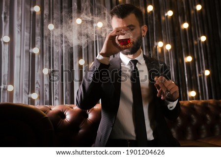 Portrait of a brutal beard man with tattoo smoking cigar in hot elegant suit  drinking whiskey alcohol in loft apartment . Couple love story concept