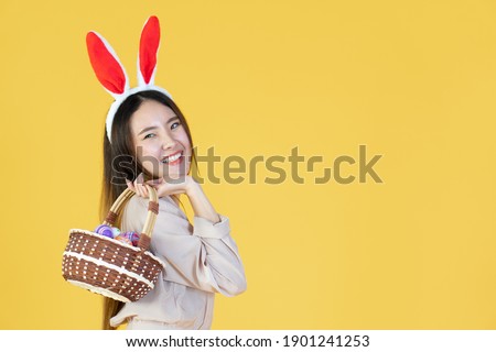 portrait of beautiful young asian woman with smiling face wearing bunny ear, holding multicolor set of easter eggs on the yellow background
