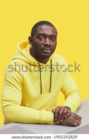 Vertical portrait of contemporary African-American man wearing hoodie and looking at camera while sitting at desk against illuminating yellow background