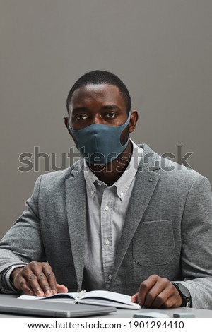 Vertical portrait of confident African-American businessman wearing mask and looking at camera while sitting at desk against minimal gray background, copy space