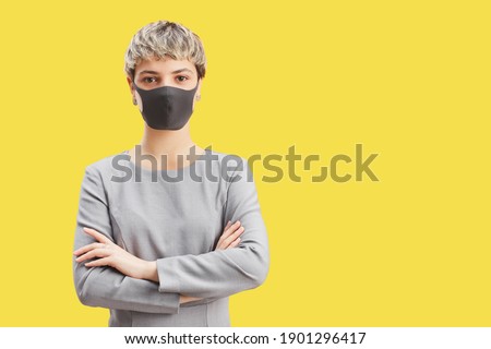 Waist up portrait of confident short-haired woman wearing mask and looking at camera while standing with arms crossed against pop yellow background, copy space
