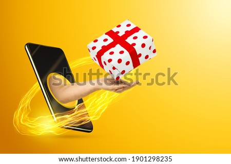 The hand crawling out of the smartphone screen holds a gift, yellow background. Christmas app for smartphones. Merry Christmas and Happy New Year concept. Copy space