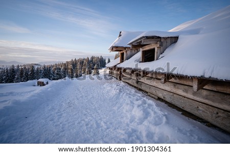 Snow covered wooden cabin facing the snowy peaks of mountains in the cold winter. Winter mountain landscape with wooden house on sunny clear day. 
