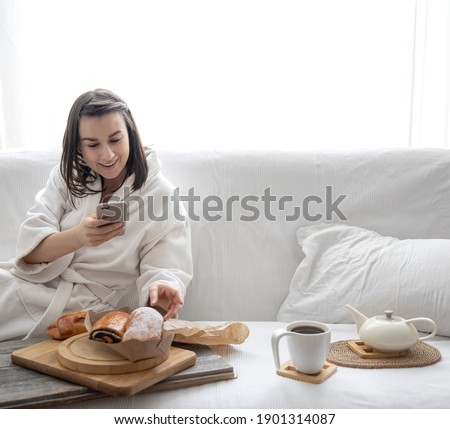 A cute young woman sits on the sofa in a robe and takes pictures of buns and coffee. Brunch and weekend concept.