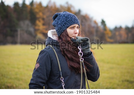 young dog breeder walk her dog. Detail on attention of a brunette who whistles disobedient dog. Woman aged 20-24 holds whistle and watches bitch. Candid portrait in authentic setting in grey moss tone Royalty-Free Stock Photo #1901325484