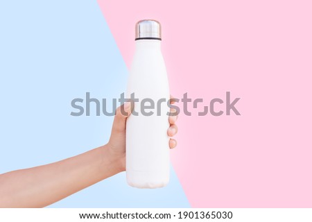 Close-up of female hand holding white reusable steel thermo water bottle isolated on two backgrounds of blue and pink colors. Royalty-Free Stock Photo #1901365030