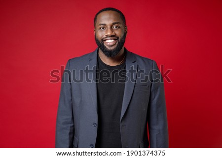 Smiling attractive confident successful young african american business man 20s wearing classic jacket suit standing and looking camera isolated on bright red color wall background studio portrait