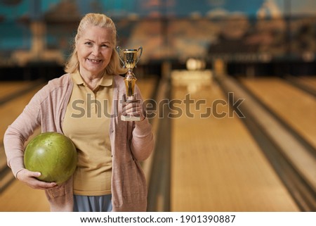 Waist up portrait of smiling mature woman holding trophy and looking at camera while standing in bowling alley after winning match, copy space