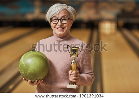Waist up portrait of smiling senior woman holding trophy and looking at camera while standing in bowling alley after winning match, copy space