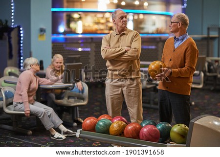 Portrait of two senior men playing bowling together with friends in background while enjoying active entertainment at bowling alley, copy space