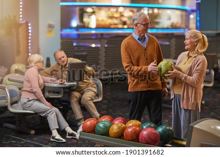 Portrait of smiling senior couple playing bowling together with friends in background while enjoying active entertainment at bowling alley, copy space