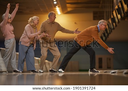 Full length side view at group of excited senior people playing bowling while enjoying active entertainment at bowling alley, copy space