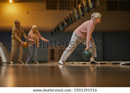 Wide angle side view at group of senior people playing bowling while enjoying active entertainment at bowling alley, copy space
