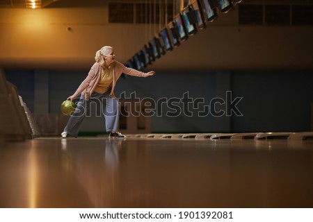 Wide angle side view at senior woman playing bowling alone while enjoying active entertainment at bowling alley, copy space
